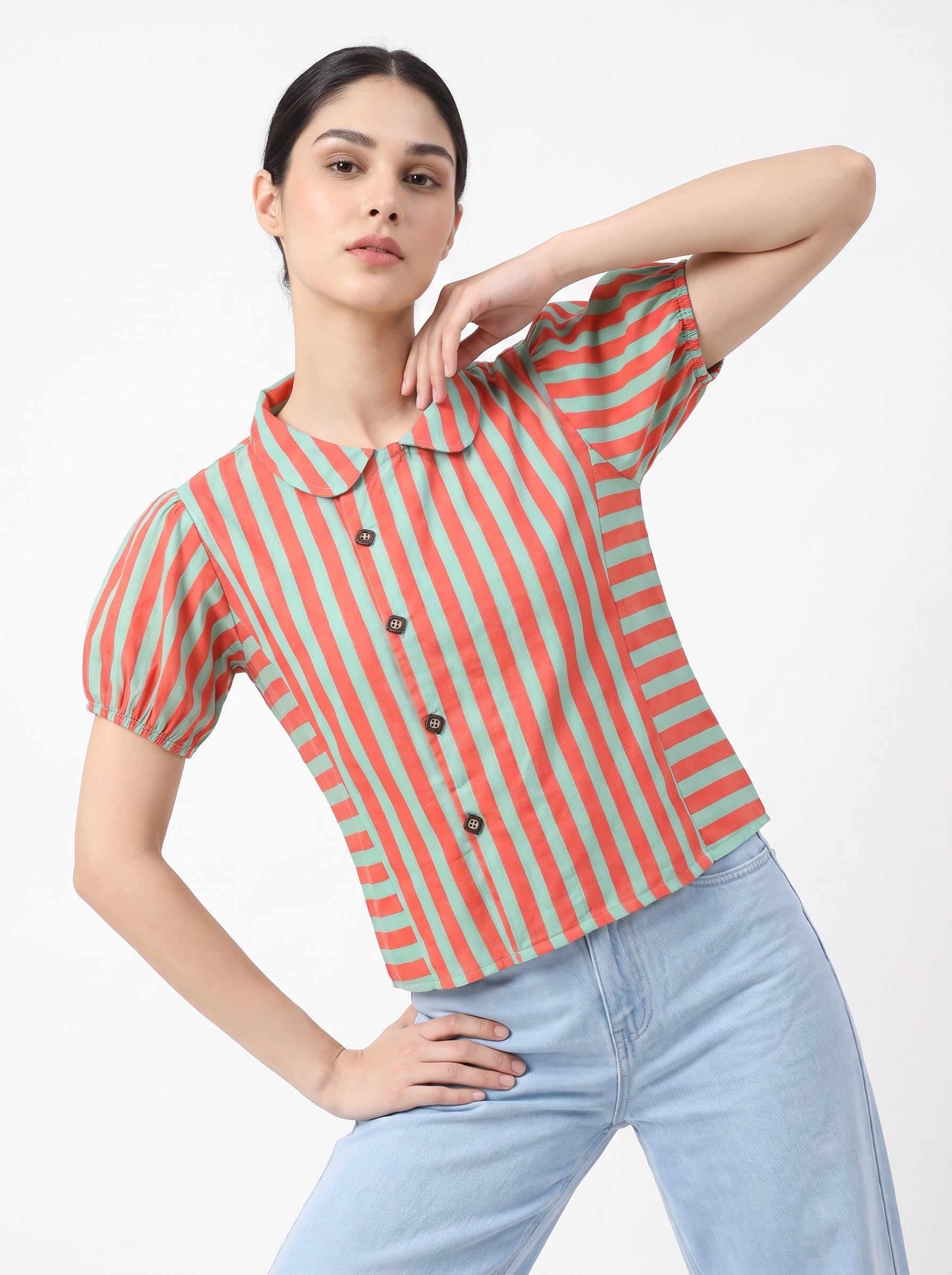 A woman modeling a short-sleeve collared button-front top with vertical coral and mint green stripes, paired with light blue jeans, against a plain light gray studio background.