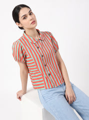A woman wearing a short-sleeve collared button-up shirt with vertical coral pink and mint green stripes, paired with light blue jeans, sitting on a white block against a white studio background.