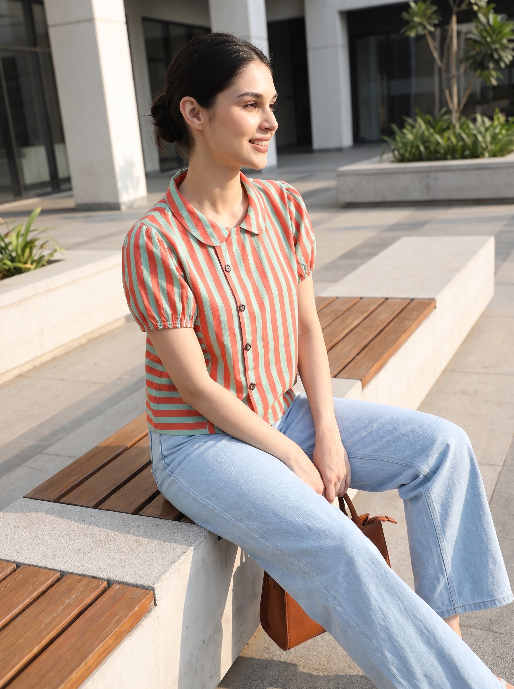 A woman sitting on a wooden bench in an outdoor plaza, wearing a short-sleeve button-up shirt with vertical orange and white stripes, paired with light blue jeans and holding a brown leather handbag.