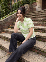 A woman sitting on outdoor stone steps wearing a light green and white vertically striped short-sleeve button-up shirt with dark gray wide-leg jeans.