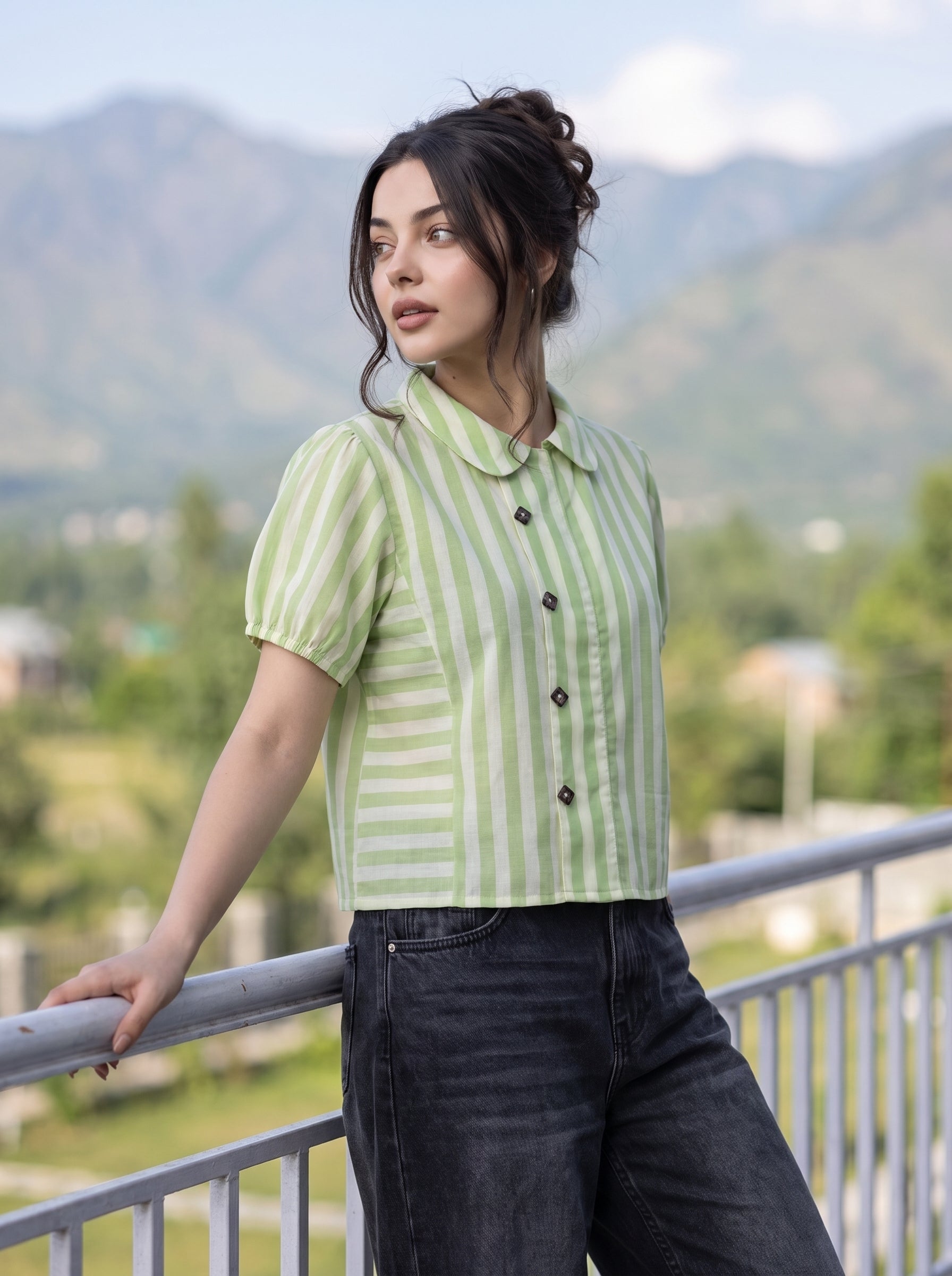 A woman wearing a light green and white vertically striped short-sleeve button-up shirt with dark jeans, leaning on a metal railing with a mountainous outdoor background.