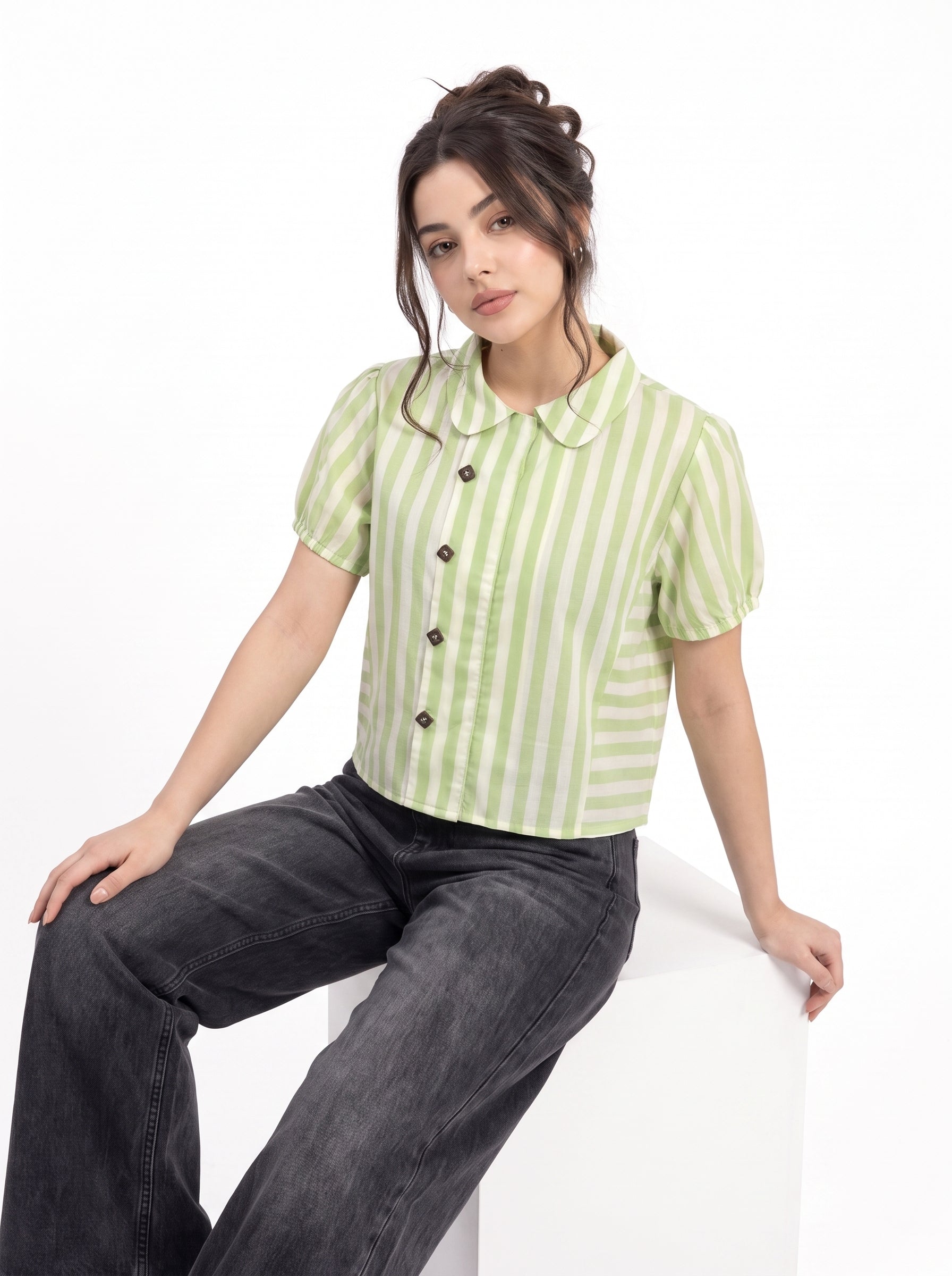 A woman wearing a light green and white vertically striped short-sleeve button-up shirt with a Peter Pan collar, paired with dark gray wide-leg jeans, sitting on a white block against a white background.