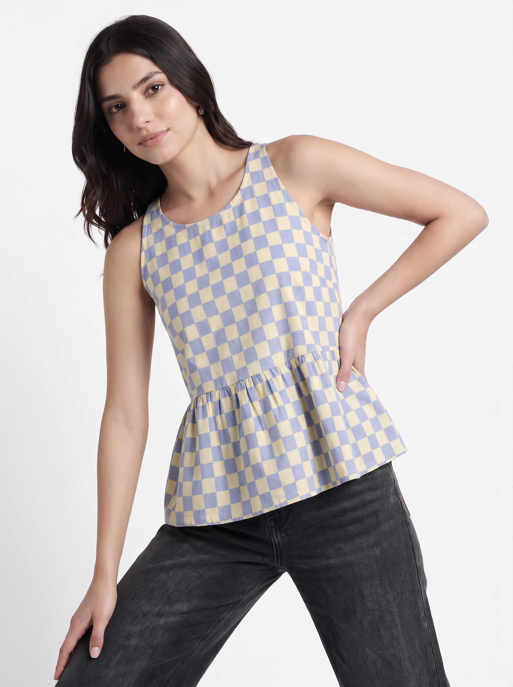 A woman modeling a sleeveless peplum top with a light blue and cream checkerboard pattern, paired with dark gray jeans, against a plain light gray background.
