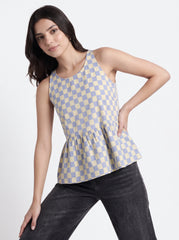 A woman modeling a sleeveless peplum top with a light blue and cream checkerboard pattern, paired with dark gray jeans, against a plain light gray background.