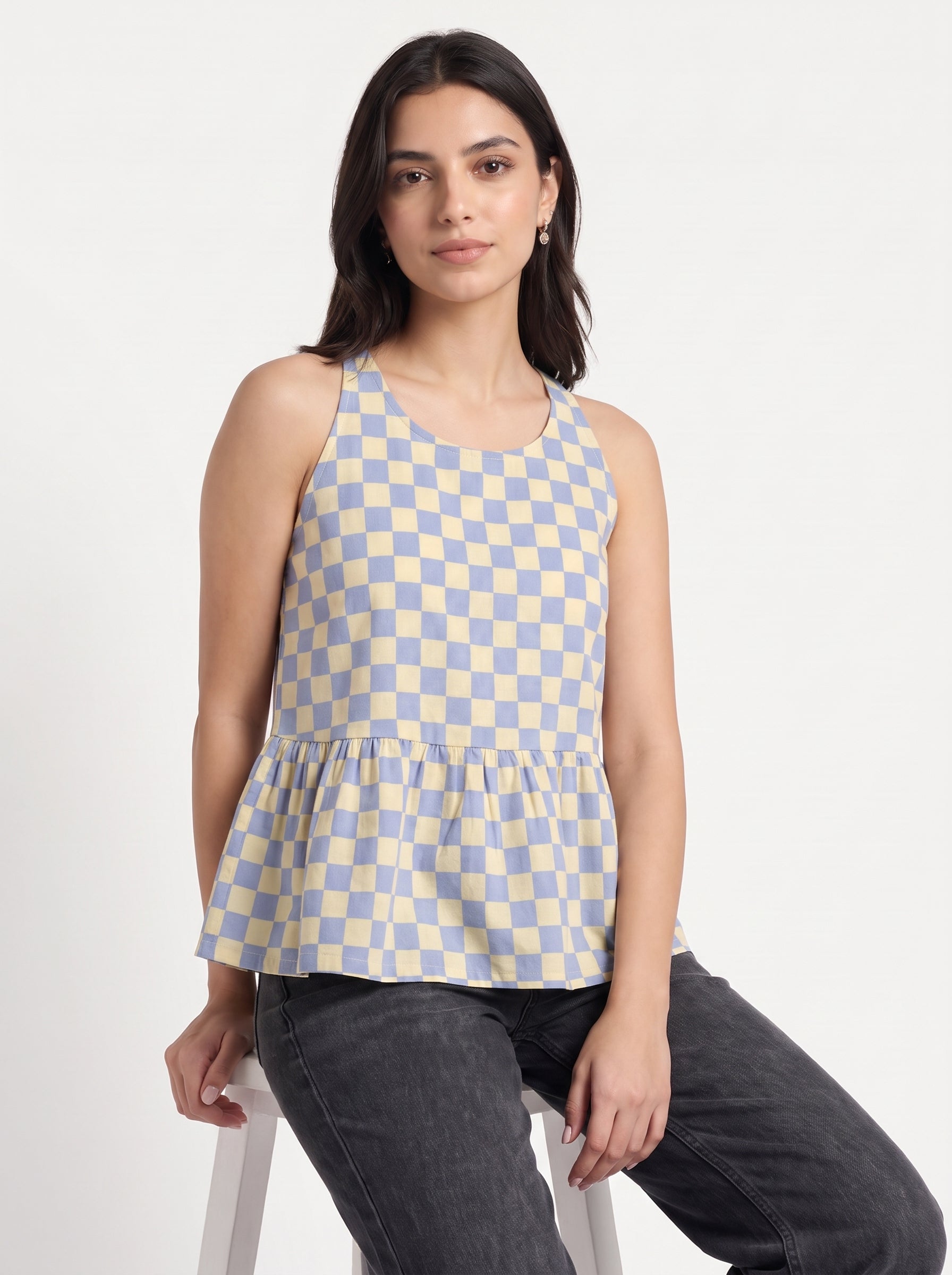 A woman wearing a sleeveless blue and beige checkered peplum top and dark gray jeans, sitting on a white stool against a plain white background.