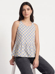 A woman wearing a sleeveless blue and beige checkered peplum top and dark gray jeans, sitting on a white stool against a plain white background.