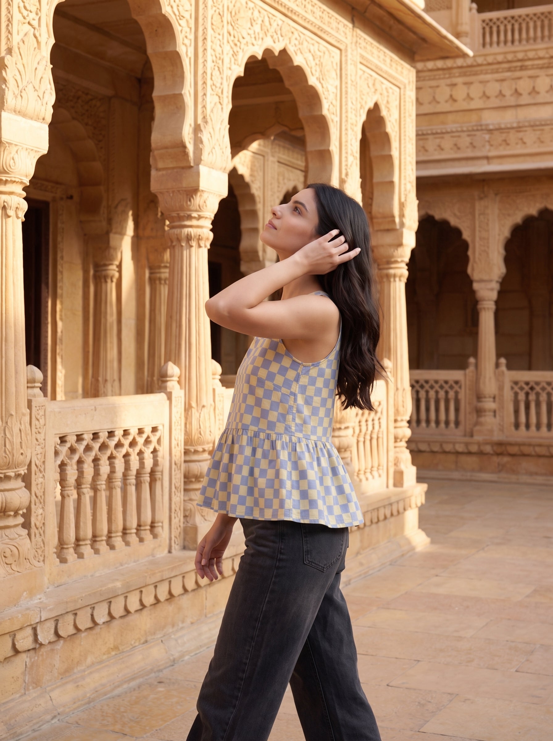 A woman wearing a light blue and beige checkered sleeveless peplum top and dark jeans walks through a sunlit courtyard with ornate sandstone arches and pillars.
