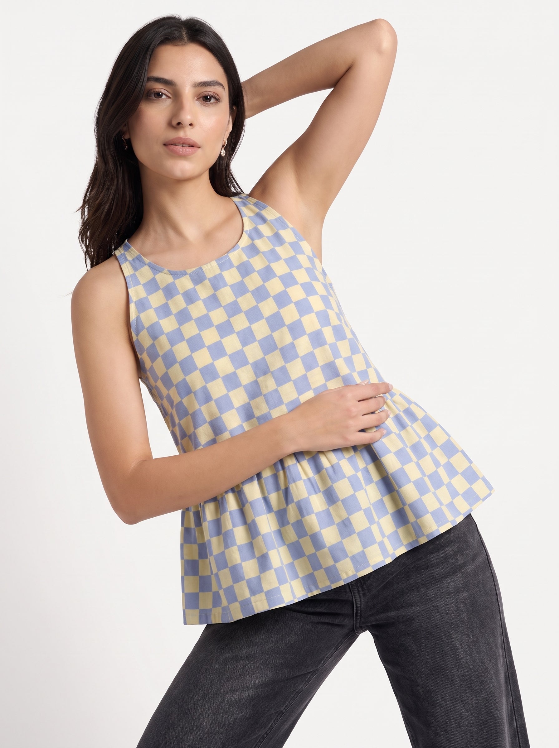 A woman modeling a sleeveless peplum top with a light blue and cream checkered pattern, paired with dark gray jeans, against a plain white studio background.