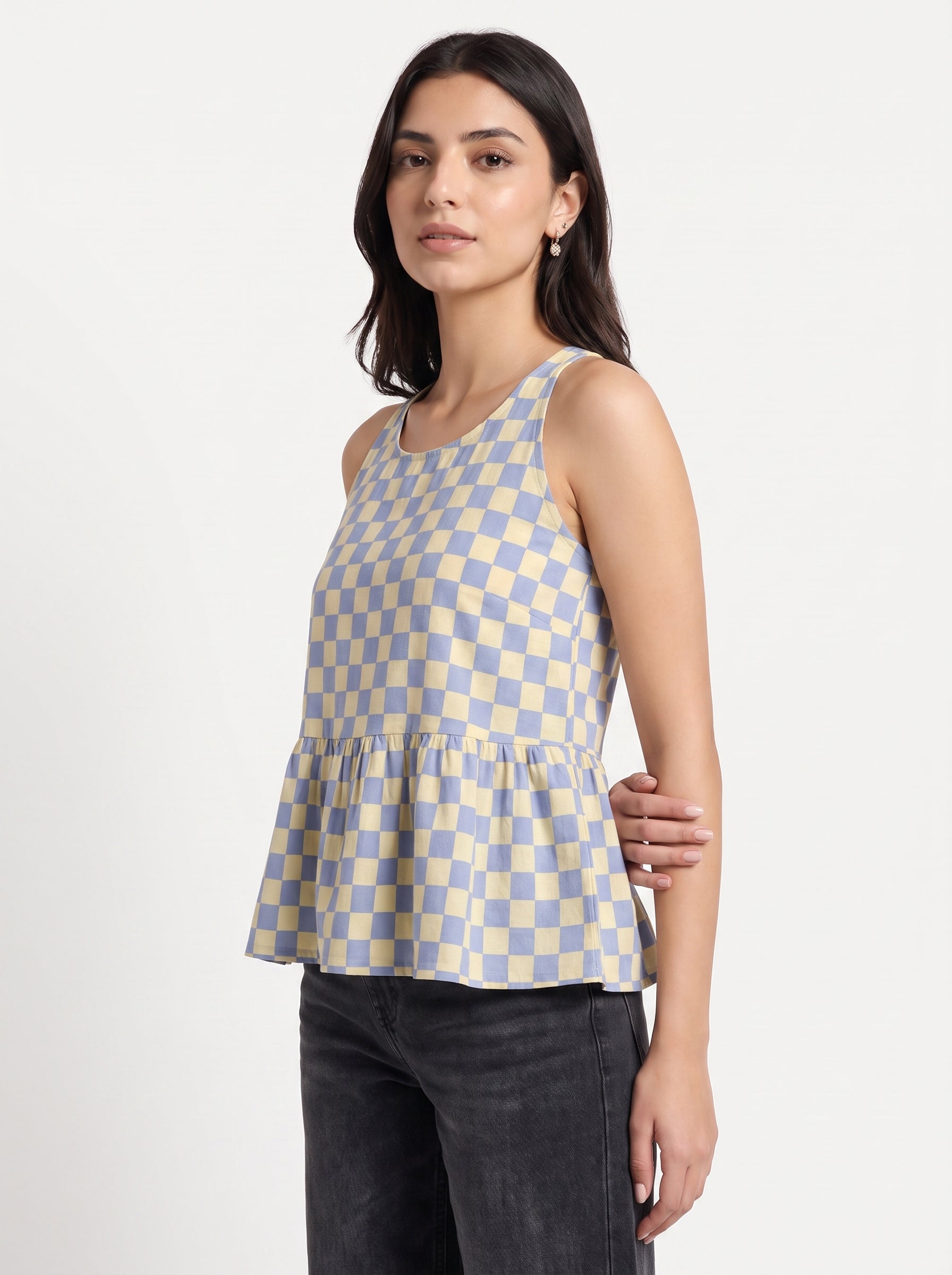 A woman modeling a sleeveless peplum top with a light blue and cream checkerboard pattern, paired with dark gray jeans, against a plain light gray studio background.