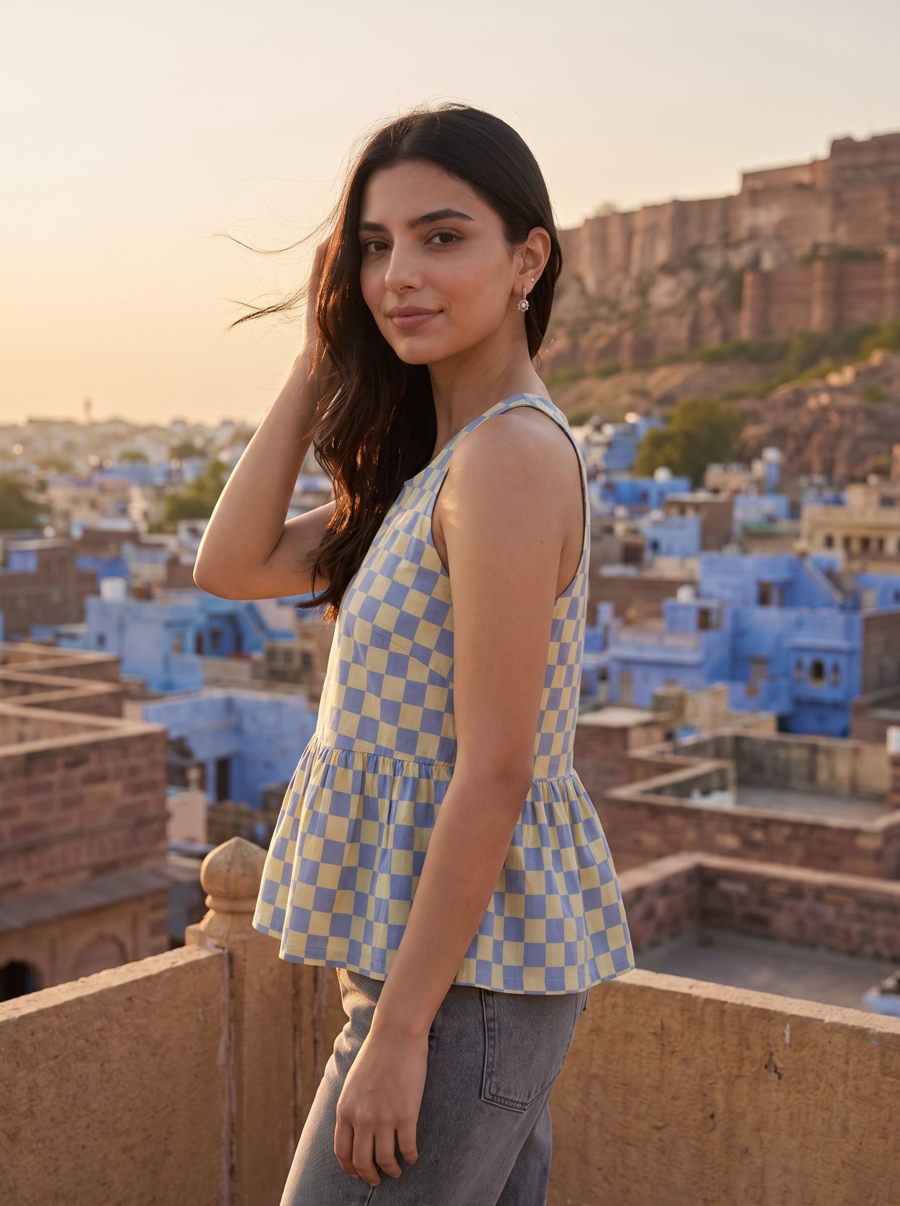 A woman wearing a light blue and white checkered sleeveless peplum top stands on a rooftop terrace with a scenic view of blue buildings and a fort in the background.