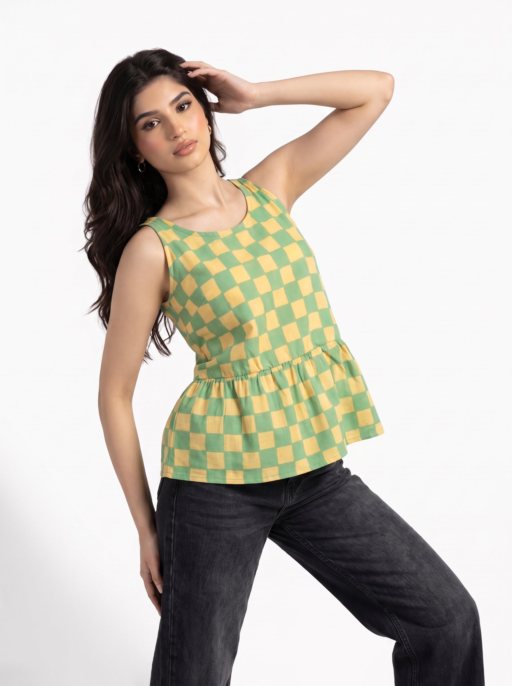 A woman modeling a sleeveless green and yellow checkered peplum top paired with dark gray jeans, posing against a plain white studio background.