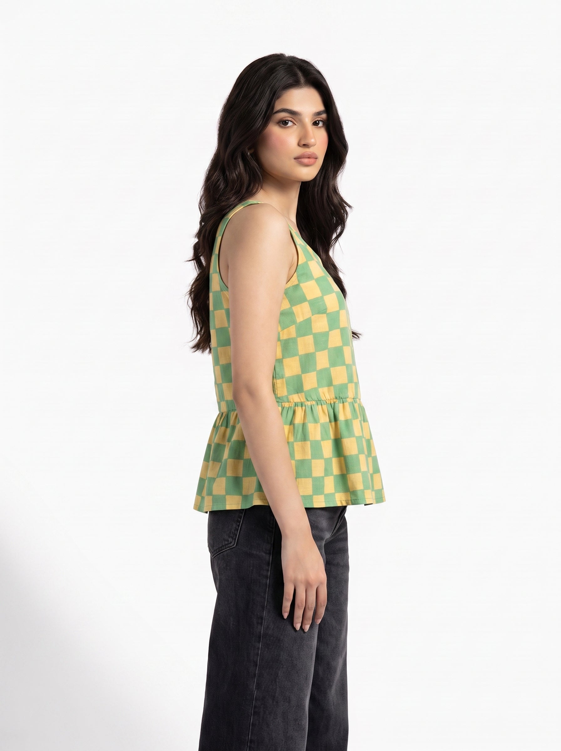 A woman modeling a sleeveless green and yellow checkered peplum top paired with dark gray jeans, shown from a side profile against a plain white studio background.