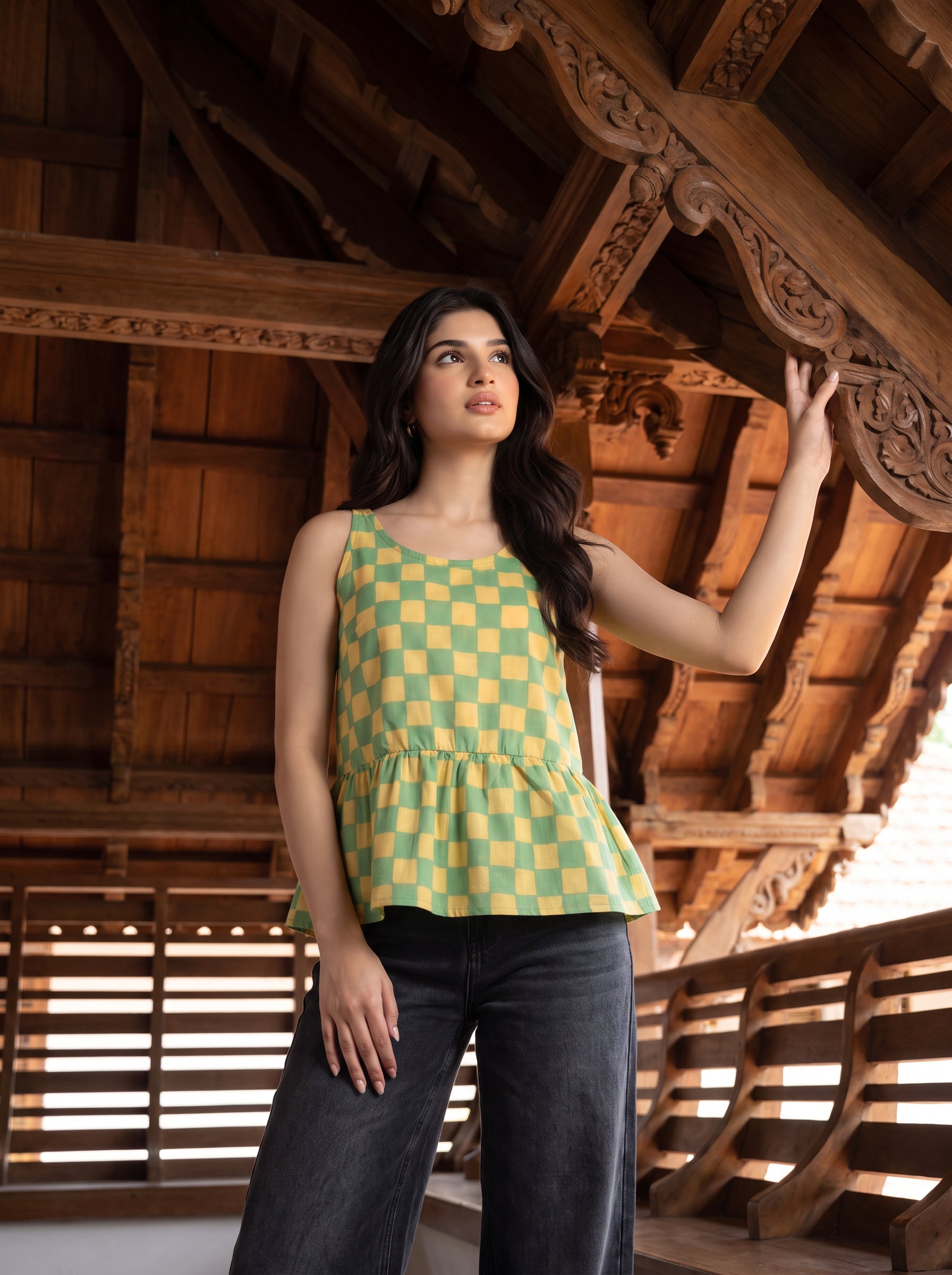 A woman wearing a green and yellow checkered sleeveless peplum top and dark jeans, posing under a traditional wooden roof structure.