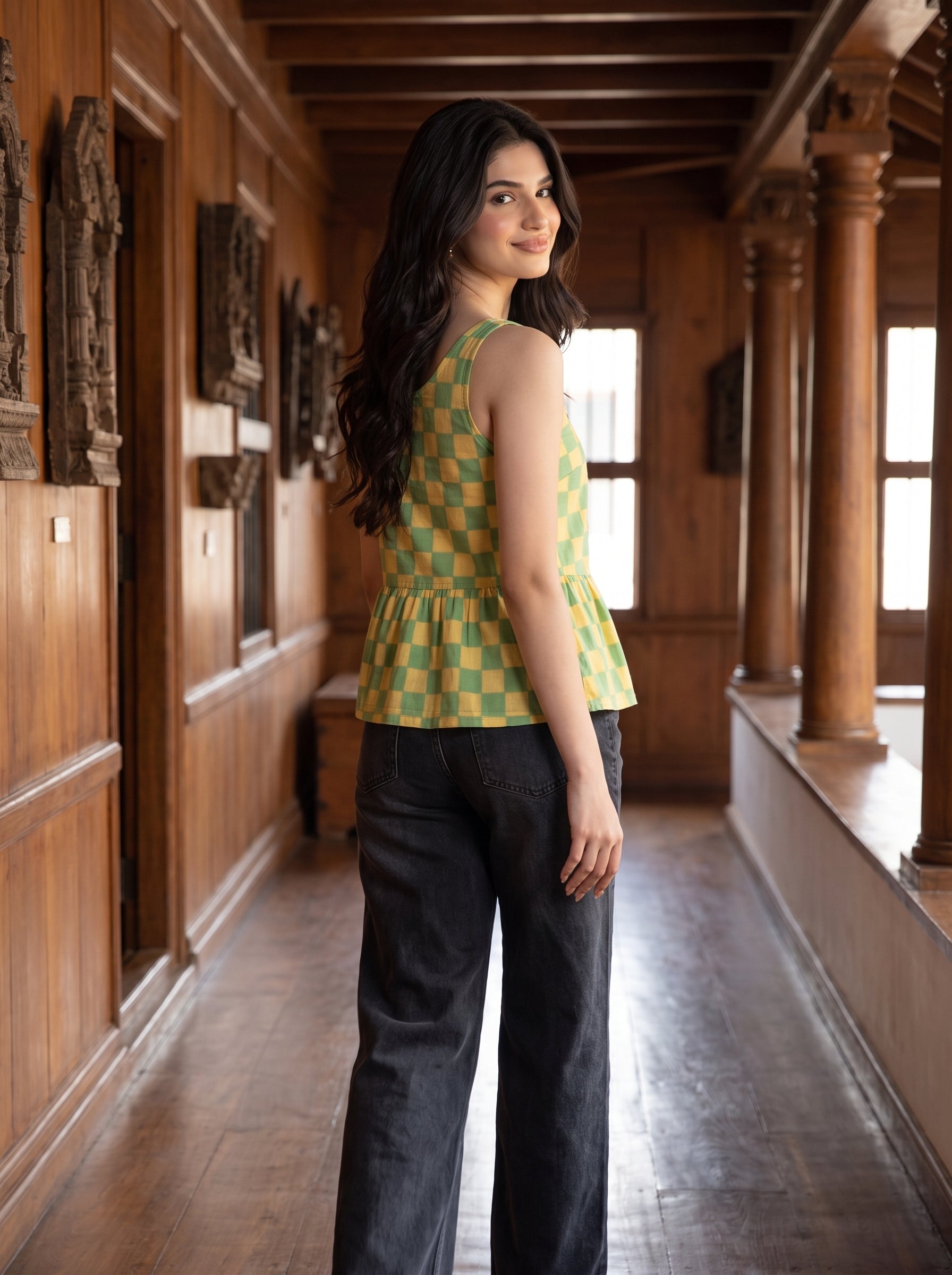 A woman stands in a wooden hallway wearing a sleeveless green and yellow checkered peplum top and dark wide-leg jeans, looking back over her shoulder.