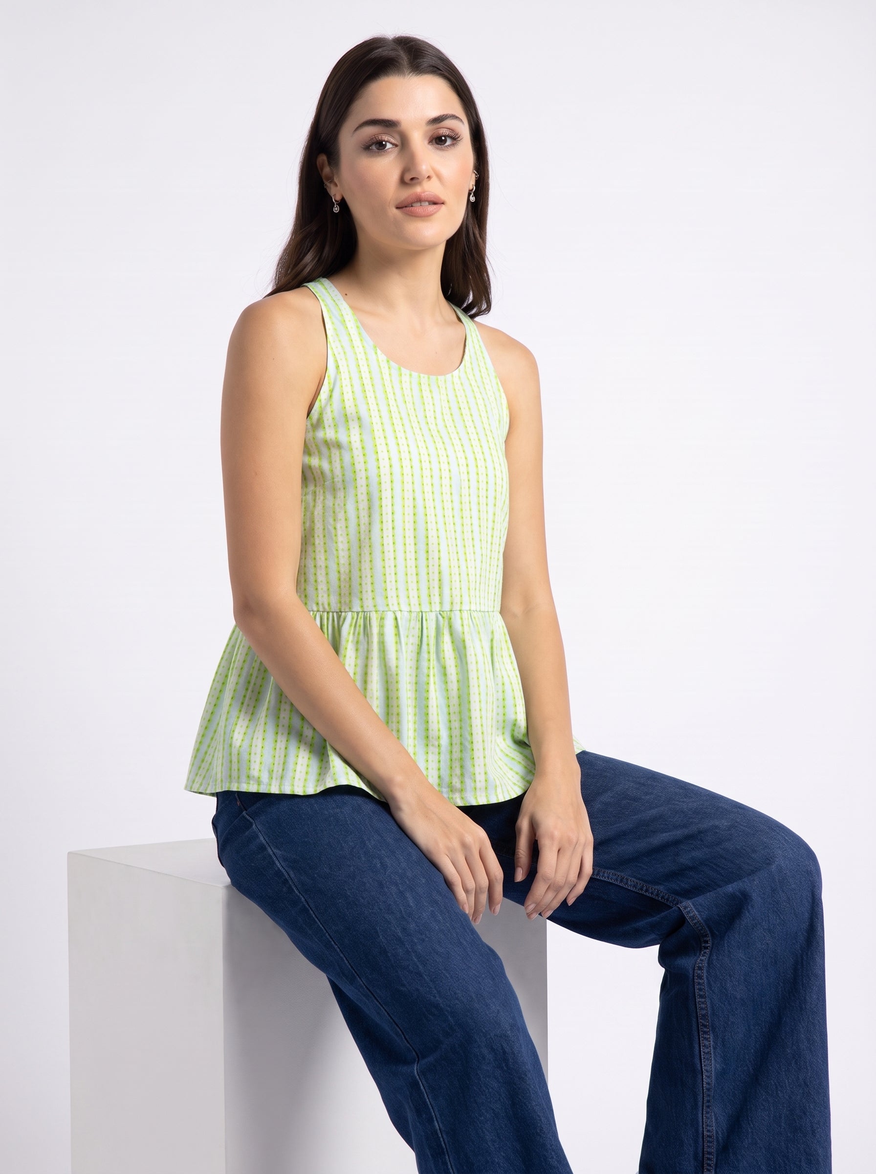 A woman wearing a light green and white gingham sleeveless peplum top and dark blue jeans, sitting on a white display block against a plain white studio background.
