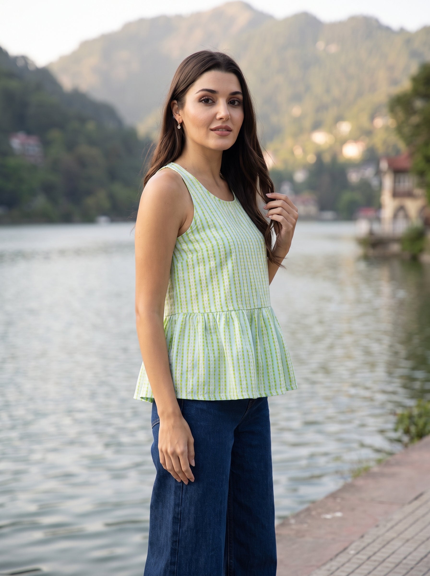 A woman stands by a lake with mountains in the background, wearing a light green and white gingham sleeveless peplum top and dark blue wide-leg pants.