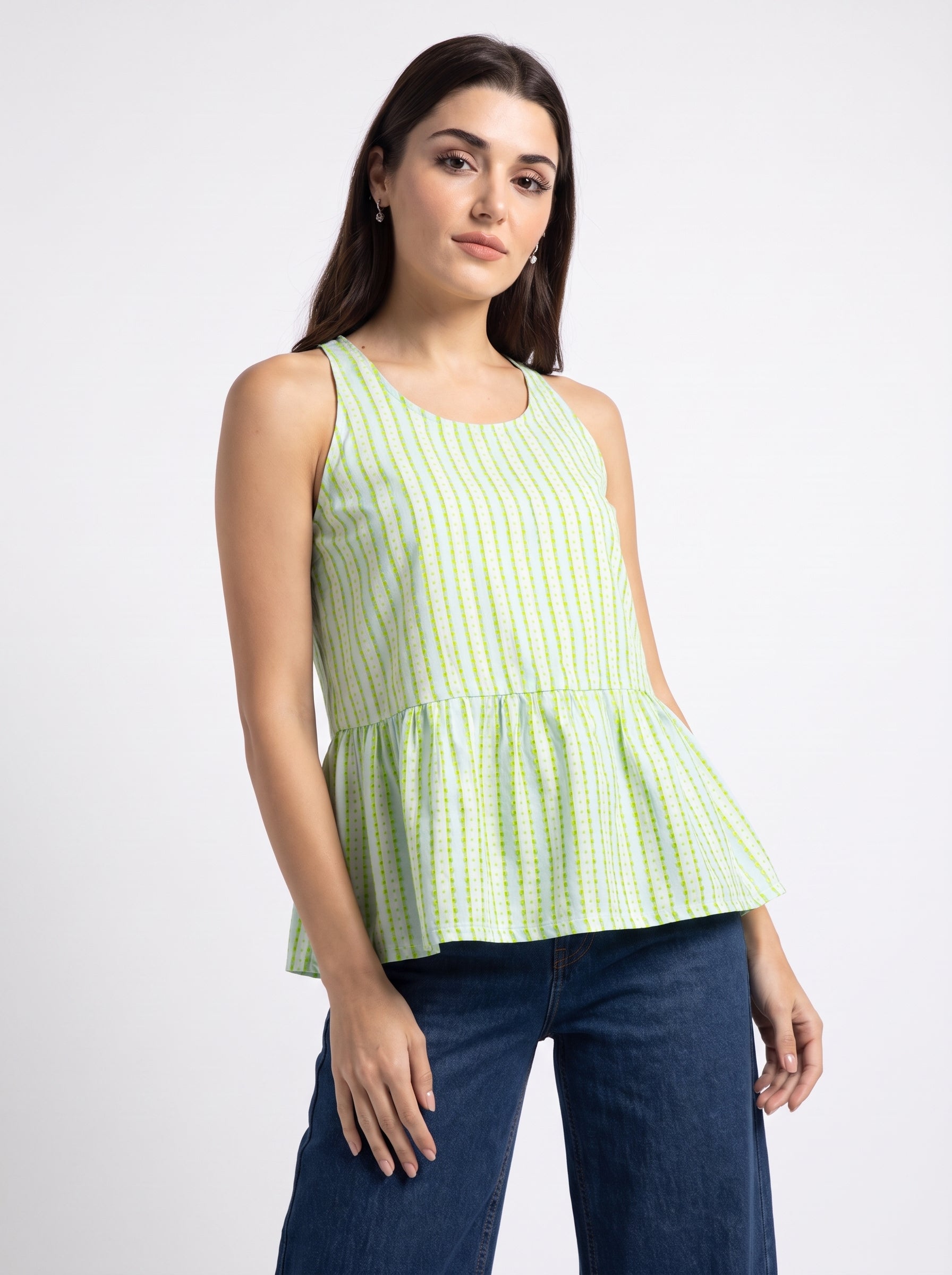 A woman modeling a sleeveless peplum top with a green and white vertical stripe pattern, paired with dark blue jeans, against a plain light gray studio background.