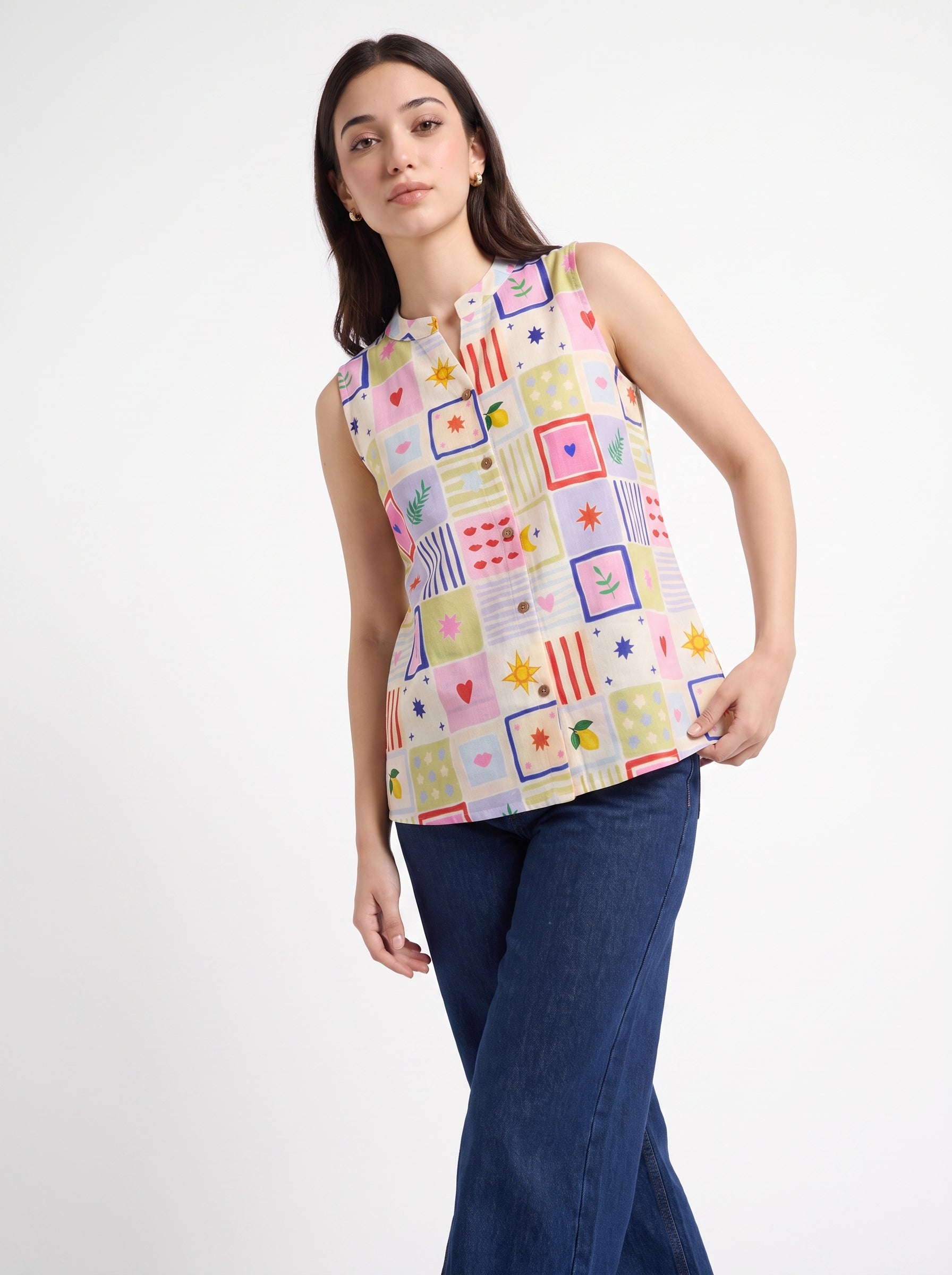 A woman modeling a sleeveless button-up top with a colorful patchwork print of geometric shapes and stars on a light pink base, paired with dark blue jeans, against a plain white studio background.