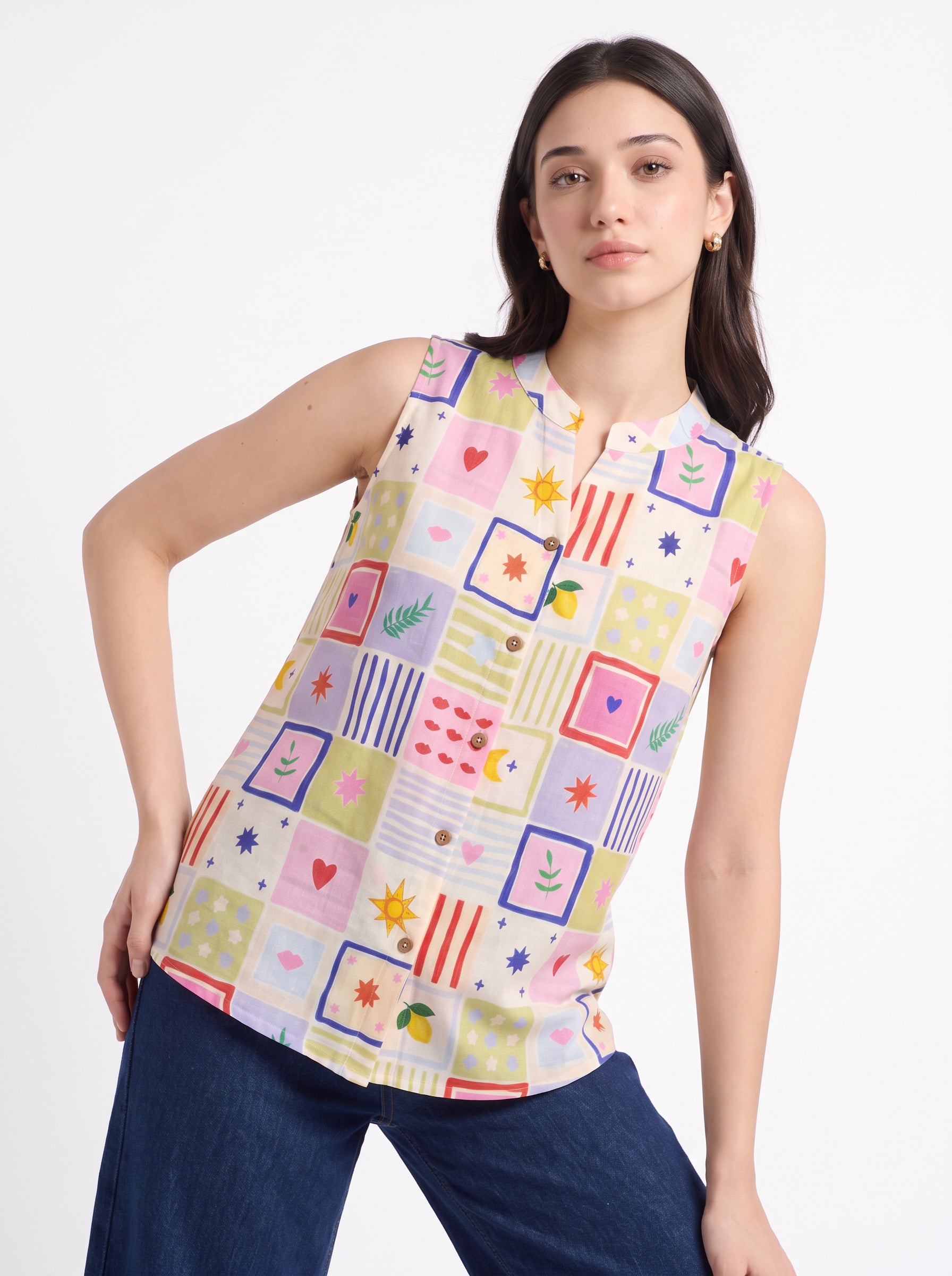A woman modeling a sleeveless button-up blouse with a colorful patchwork print of hearts, stars, stripes, and geometric shapes, paired with dark blue jeans against a plain white background.