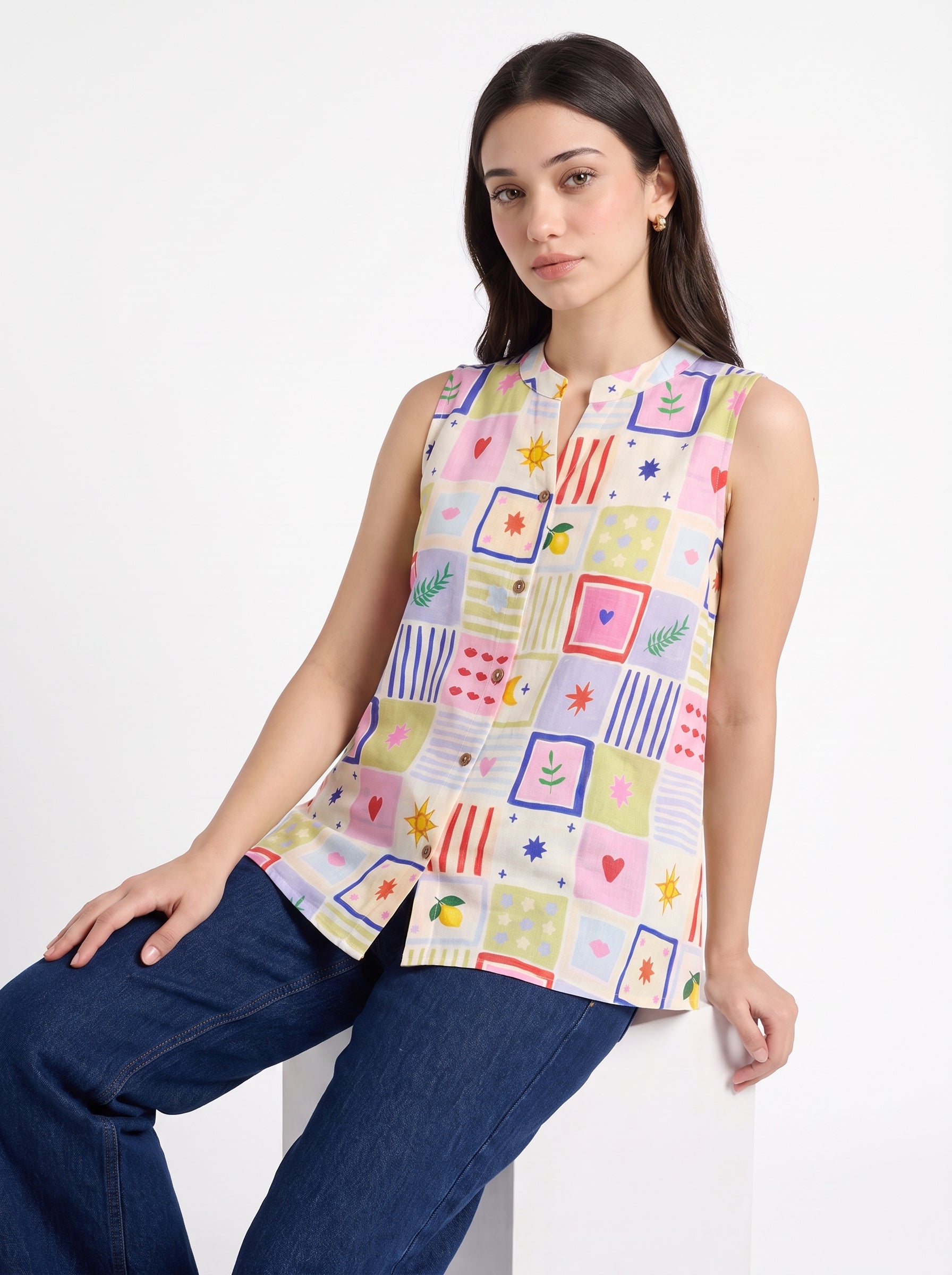 A woman wearing a sleeveless button-up top with a colorful patchwork print, paired with dark blue jeans, posing against a white studio background.