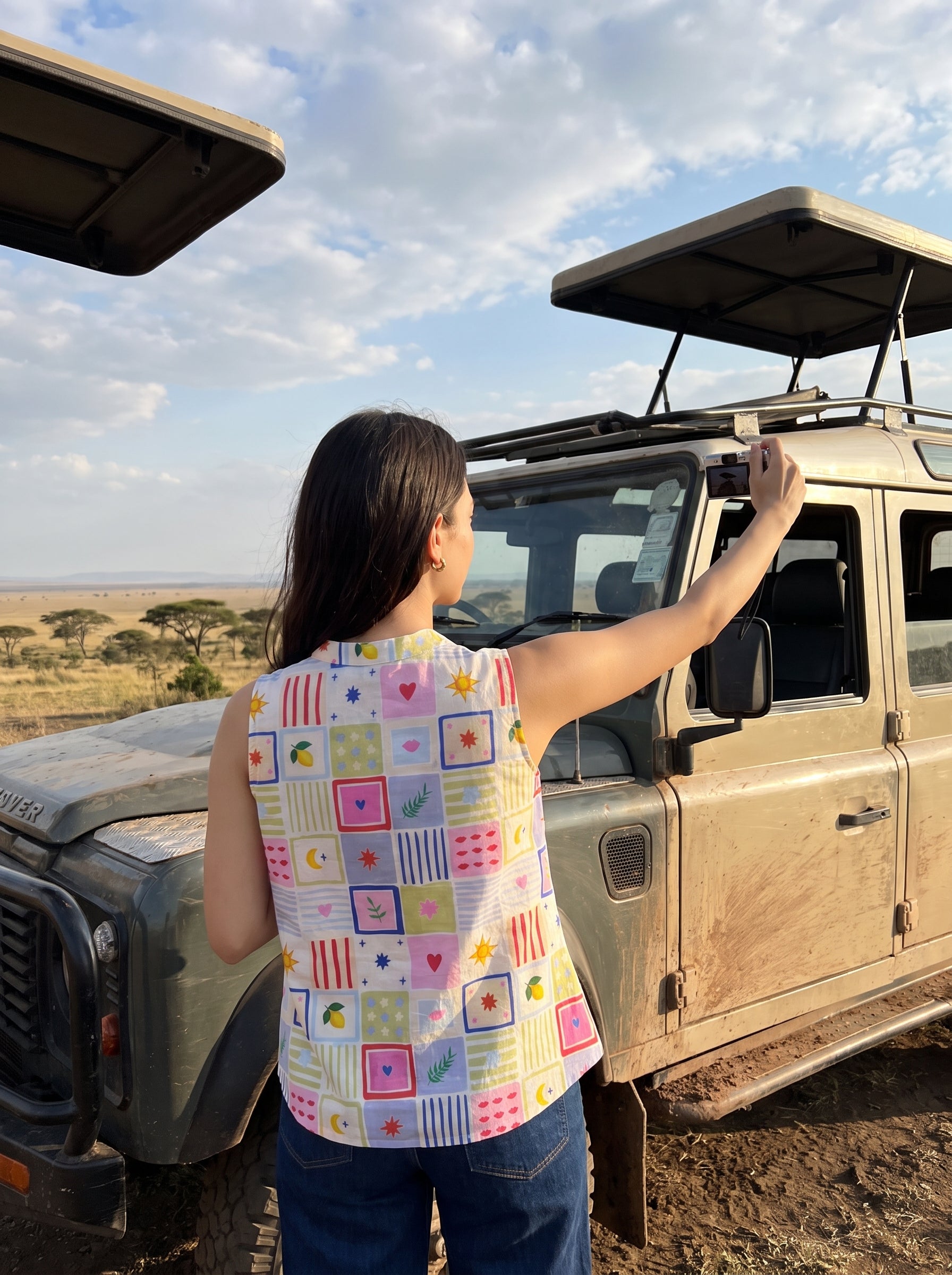 A woman stands next to a safari vehicle in a dry grassland, wearing a colorful patchwork quilt-patterned sleeveless top and blue jeans, looking out over the savanna under a cloudy sky.