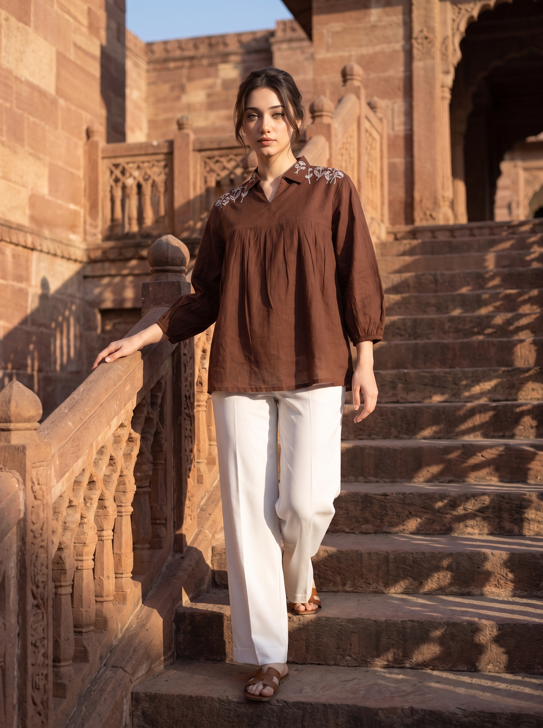 A woman wearing a brown long-sleeve blouse with embroidered shoulders and white wide-leg pants walks down stone stairs in front of a historic sandstone building.