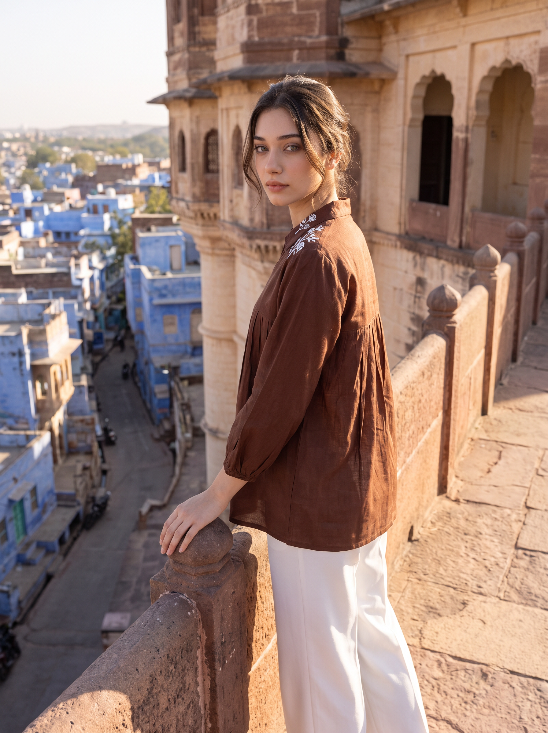 A woman stands on a stone balcony overlooking a blue-painted village, wearing a brown long-sleeve top with white floral embroidery at the collar and white wide-leg pants.