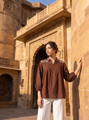 A woman stands outdoors against a sunlit sandstone architectural structure, wearing a brown long-sleeve blouse with embroidered detailing at the collar and white wide-leg pants.