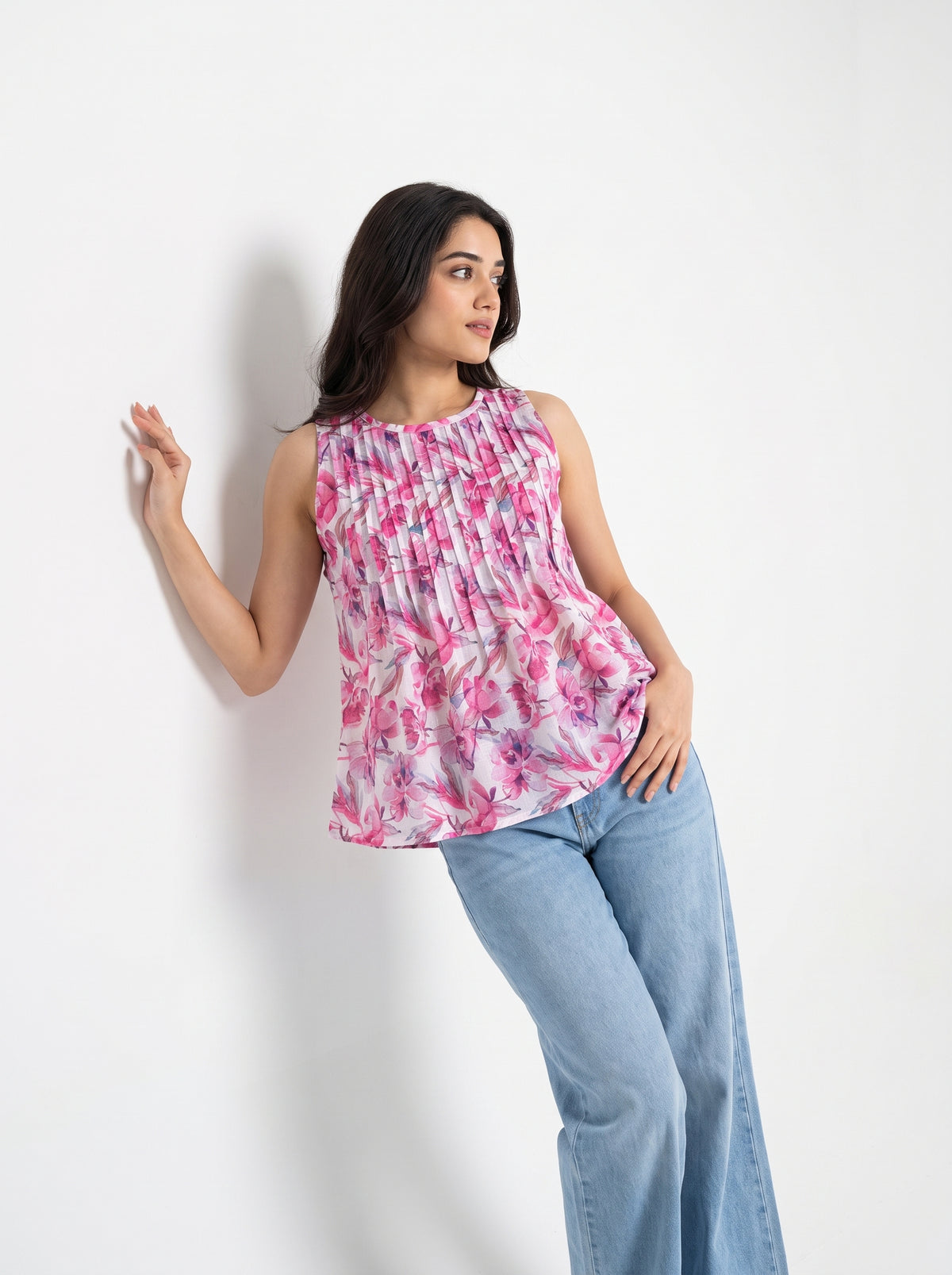 A woman wearing a sleeveless, ruffled peplum top with a pink, purple, and white floral pattern, paired with light blue jeans, posing against a plain white wall.