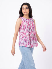 A woman modeling a sleeveless, pleated top with a pink and purple floral print on a white base, paired with light blue jeans, against a plain white background.