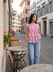 A woman stands on a cobblestone street wearing a sleeveless, smocked top with a pink and white floral pattern, paired with light blue jeans.