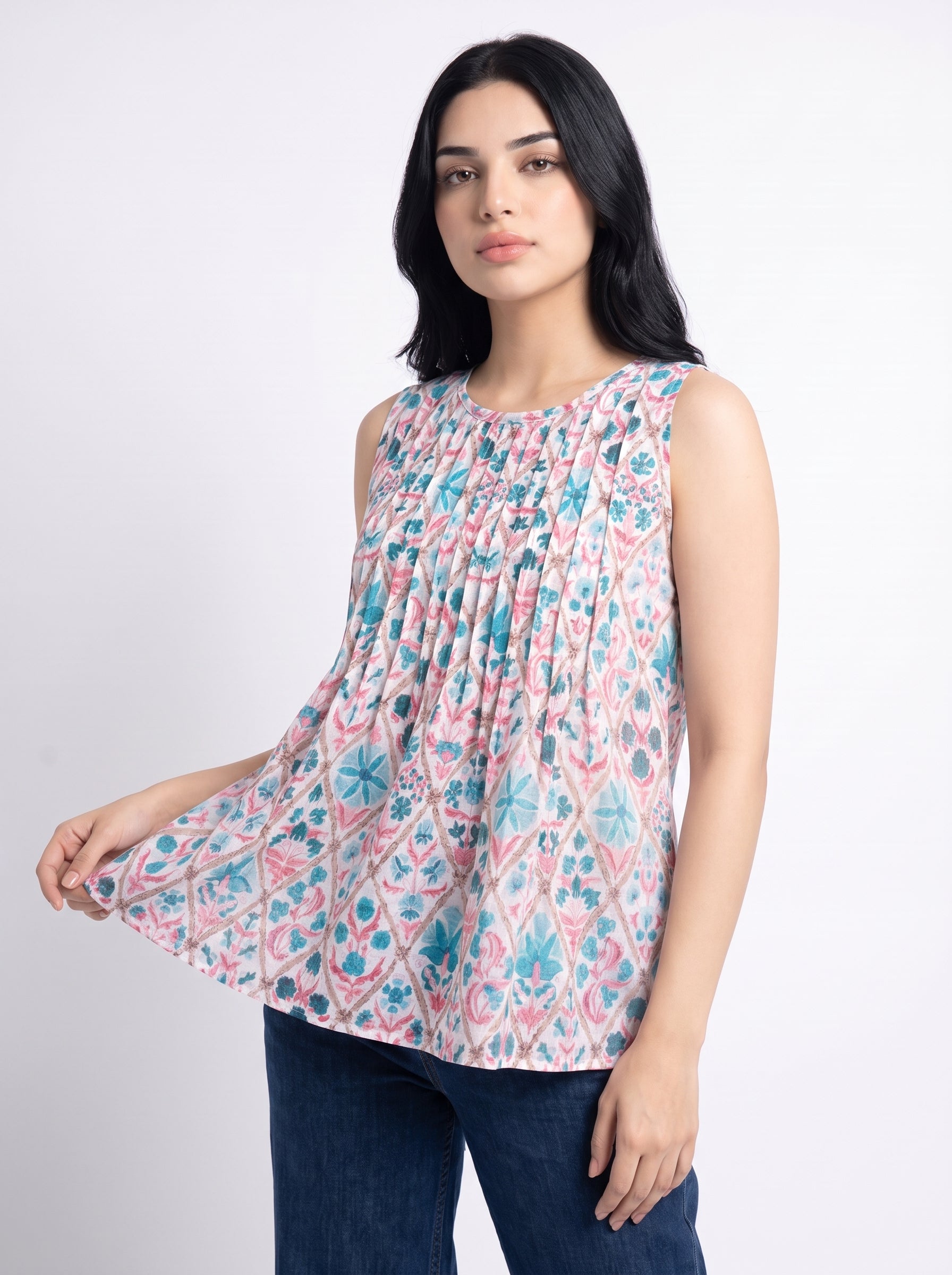 A woman modeling a sleeveless, smocked top with a pink, blue, and white geometric pattern, paired with dark blue jeans, against a plain light gray background.