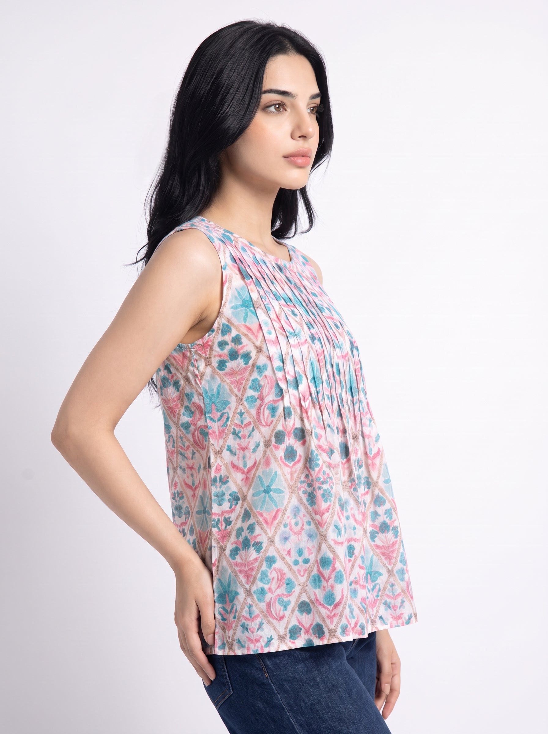 A woman wearing a sleeveless, pleated top with a pink, blue, and white geometric pattern, paired with dark blue jeans, photographed against a light gray studio background.
