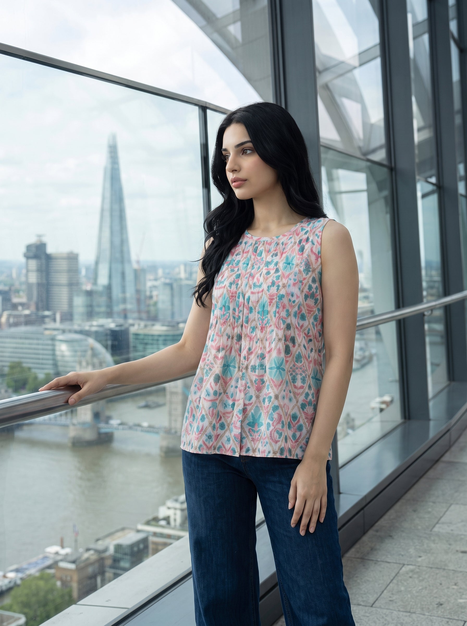 A woman stands on a balcony overlooking a city skyline with a river, wearing a sleeveless top with a pink, teal, and white abstract pattern and dark blue jeans.