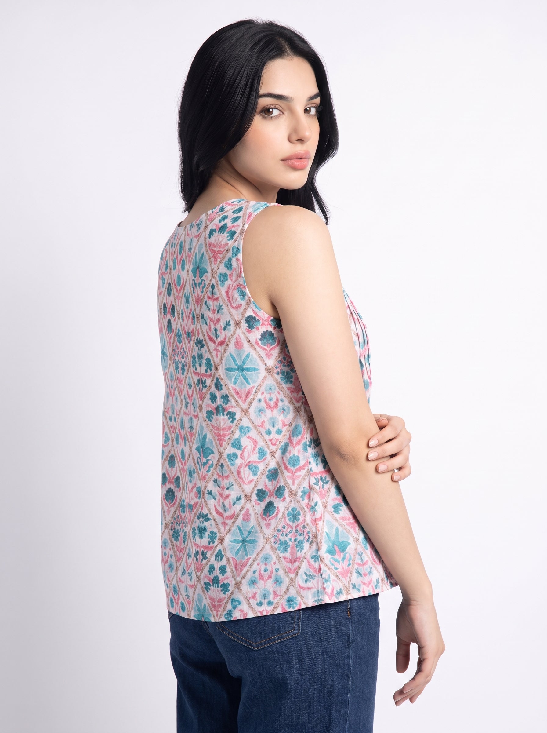 A woman shown from the back wearing a sleeveless top with a pink, blue, and white geometric diamond pattern, paired with dark blue jeans, against a light gray studio background.
