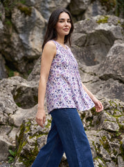 A woman wearing a sleeveless lavender and purple floral print top with blue jeans, standing outdoors in front of a rocky, mossy cliff background.