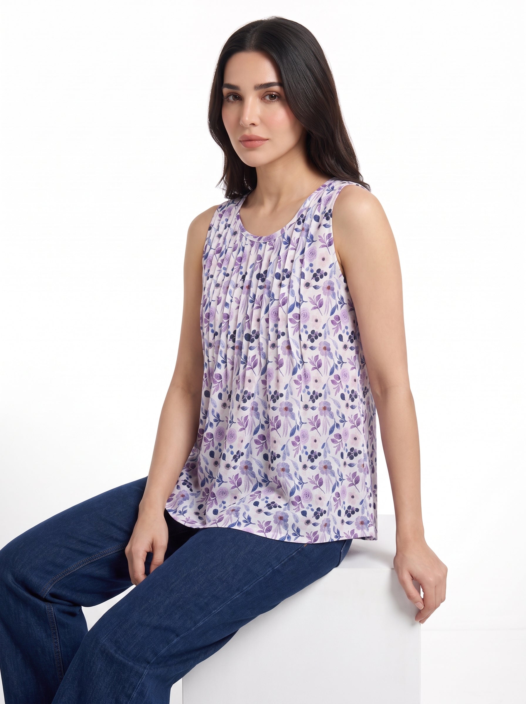 A woman wearing a sleeveless purple and white floral print top with pleated detailing, paired with dark blue jeans, seated against a plain white background.