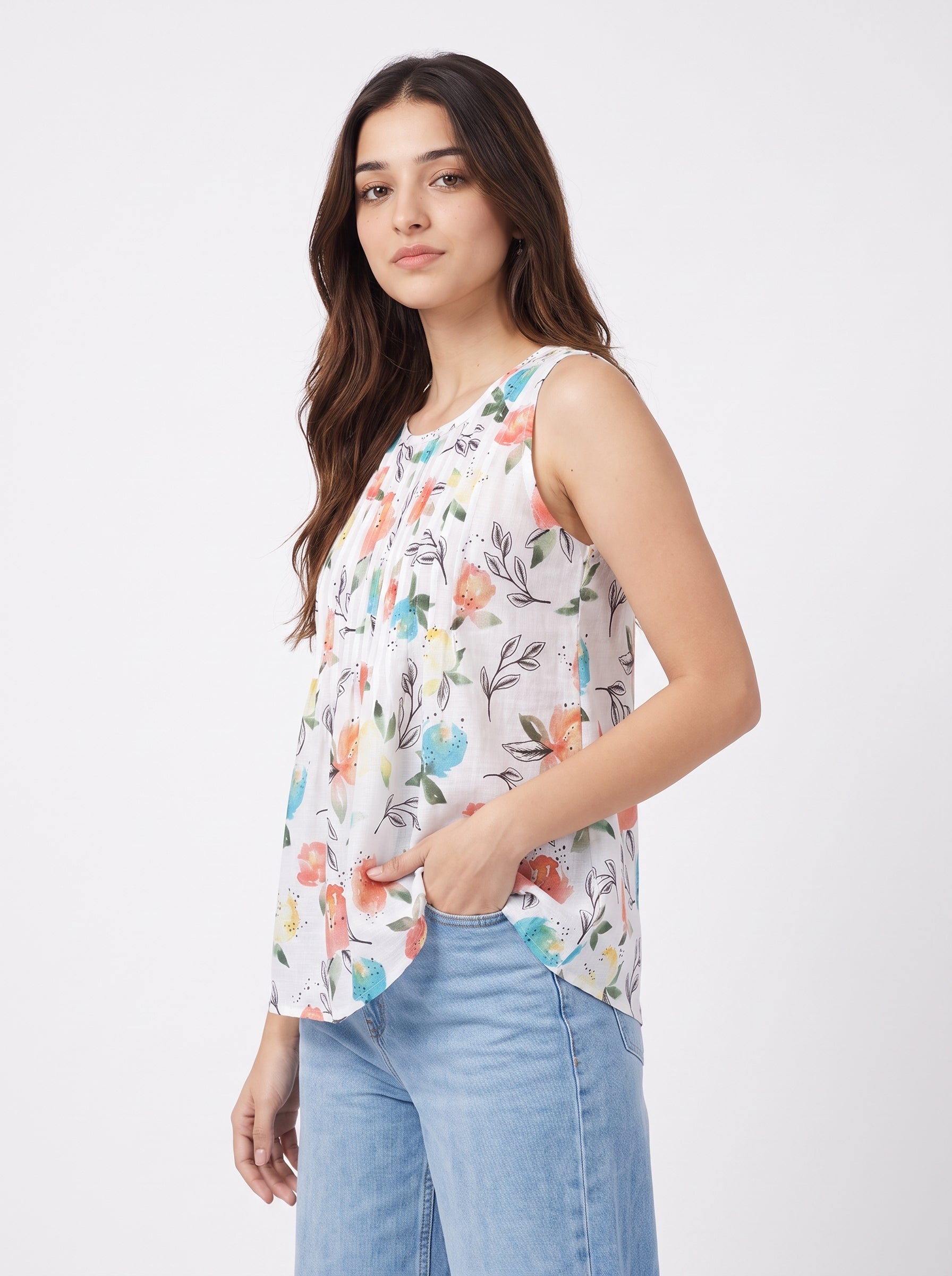 A woman wearing a sleeveless white top with a colorful floral print and light blue jeans, standing against a plain light gray studio background.