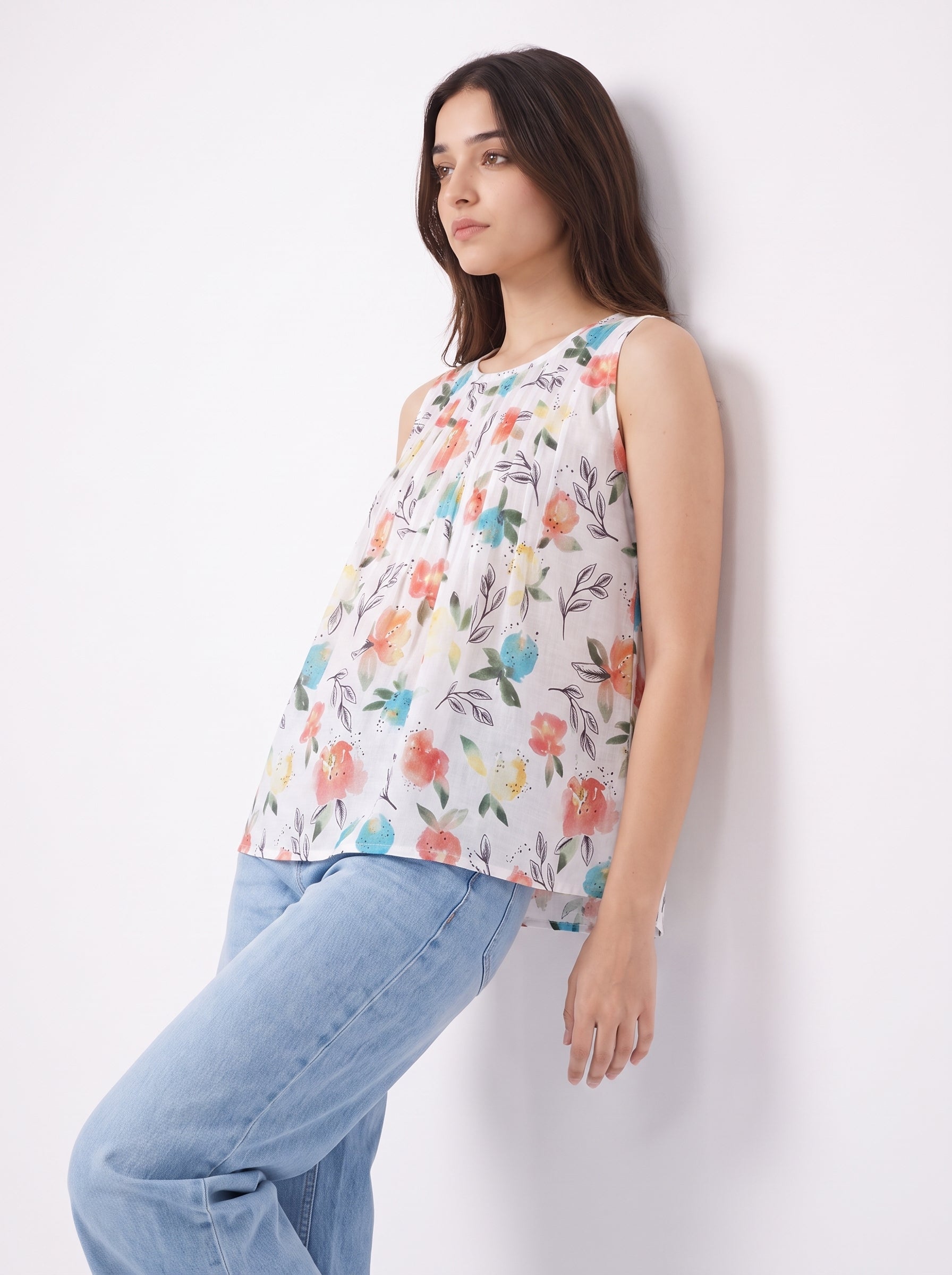 A woman wearing a sleeveless white top with a colorful floral print and light blue jeans, leaning against a plain white wall.