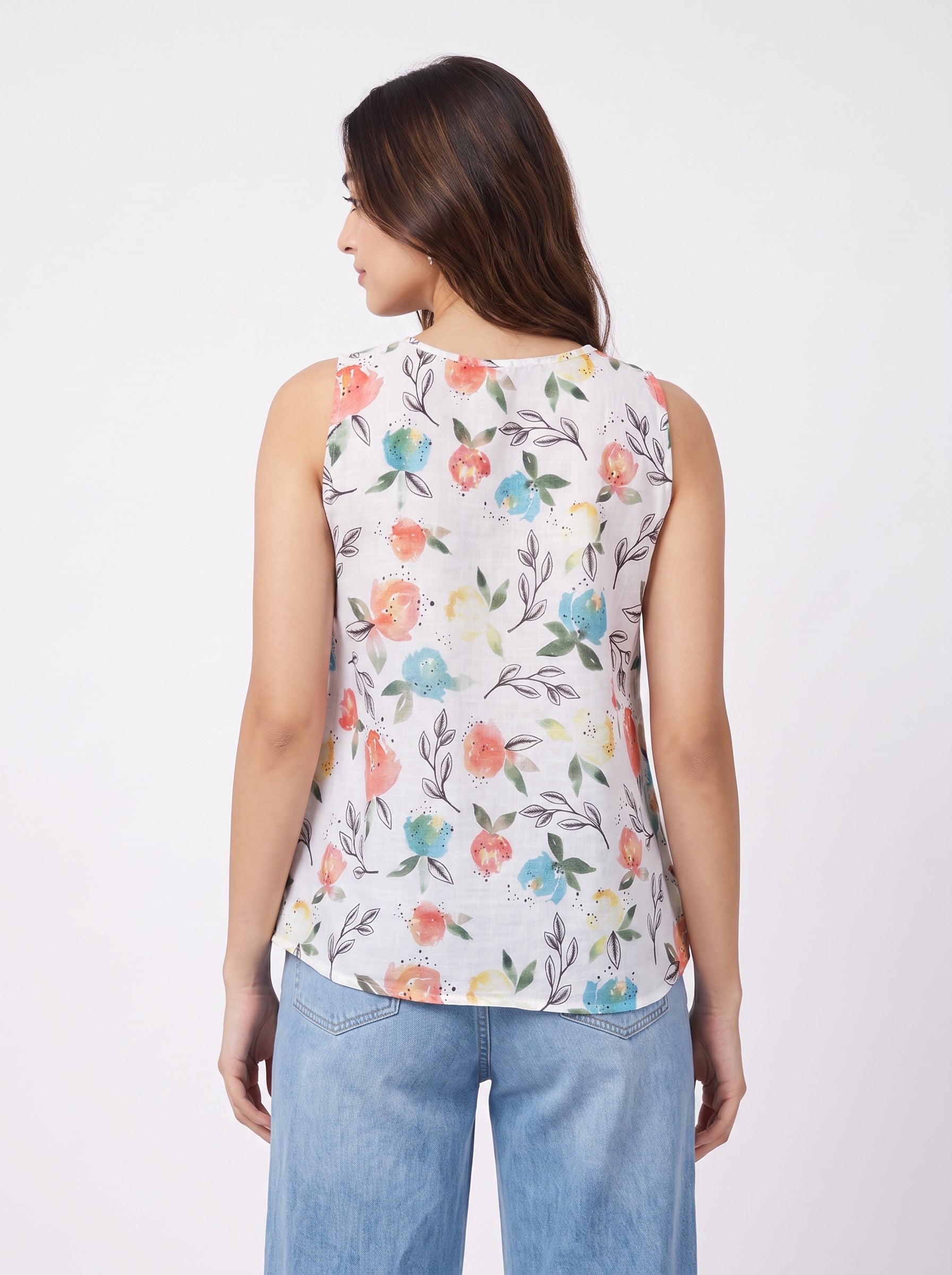 A woman shown from the back wearing a sleeveless white top with a colorful floral print and light blue jeans, against a plain light gray studio background.