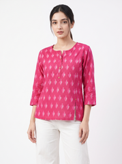 A woman modeling a bright pink three-quarter sleeve top with a white ikat pattern, paired with white pants, against a plain white studio background.