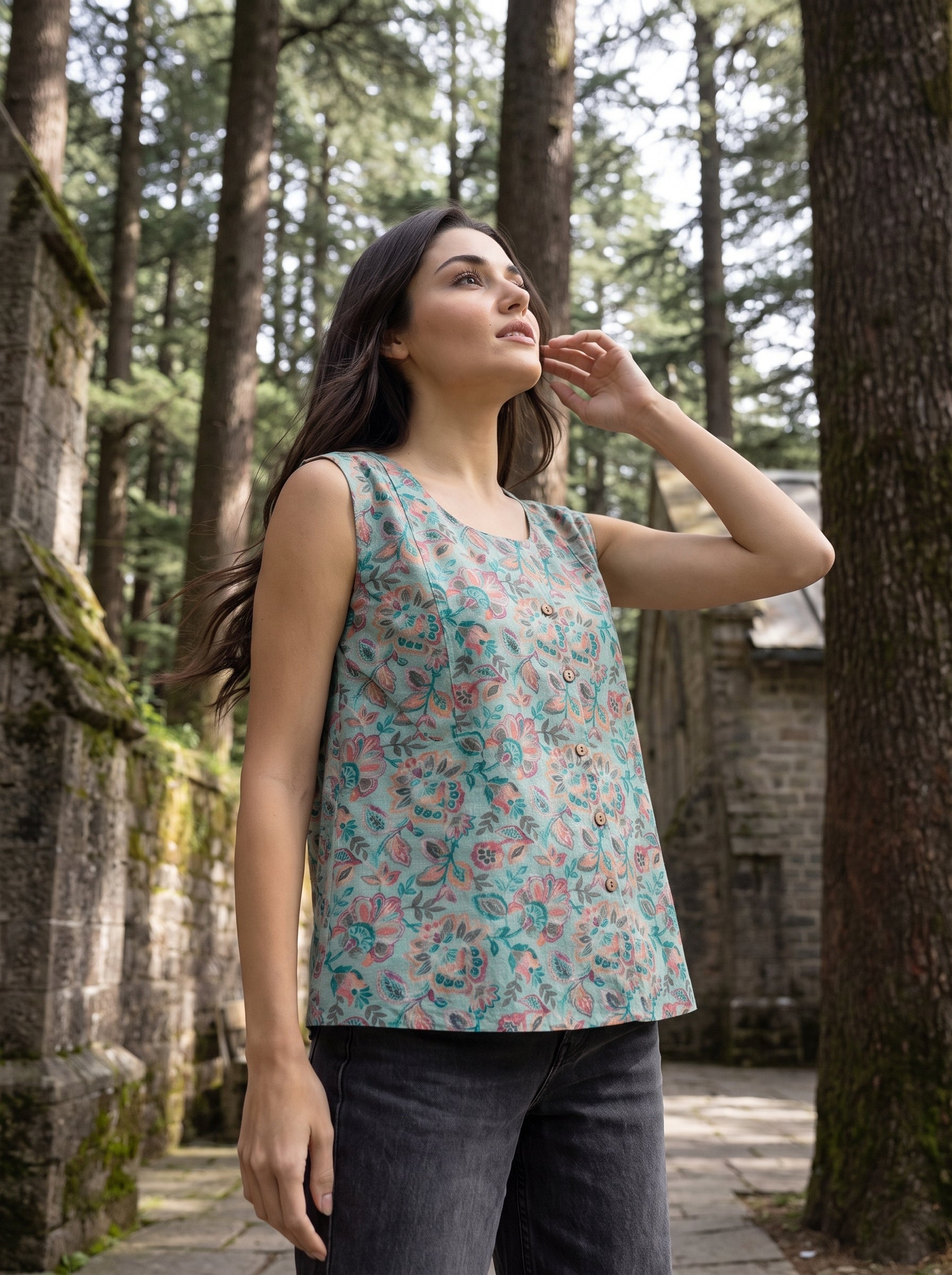 A woman wearing a sleeveless floral top and dark jeans stands outdoors in a wooded area with stone walls and tall trees. The top has a light teal base with pink and green floral print.