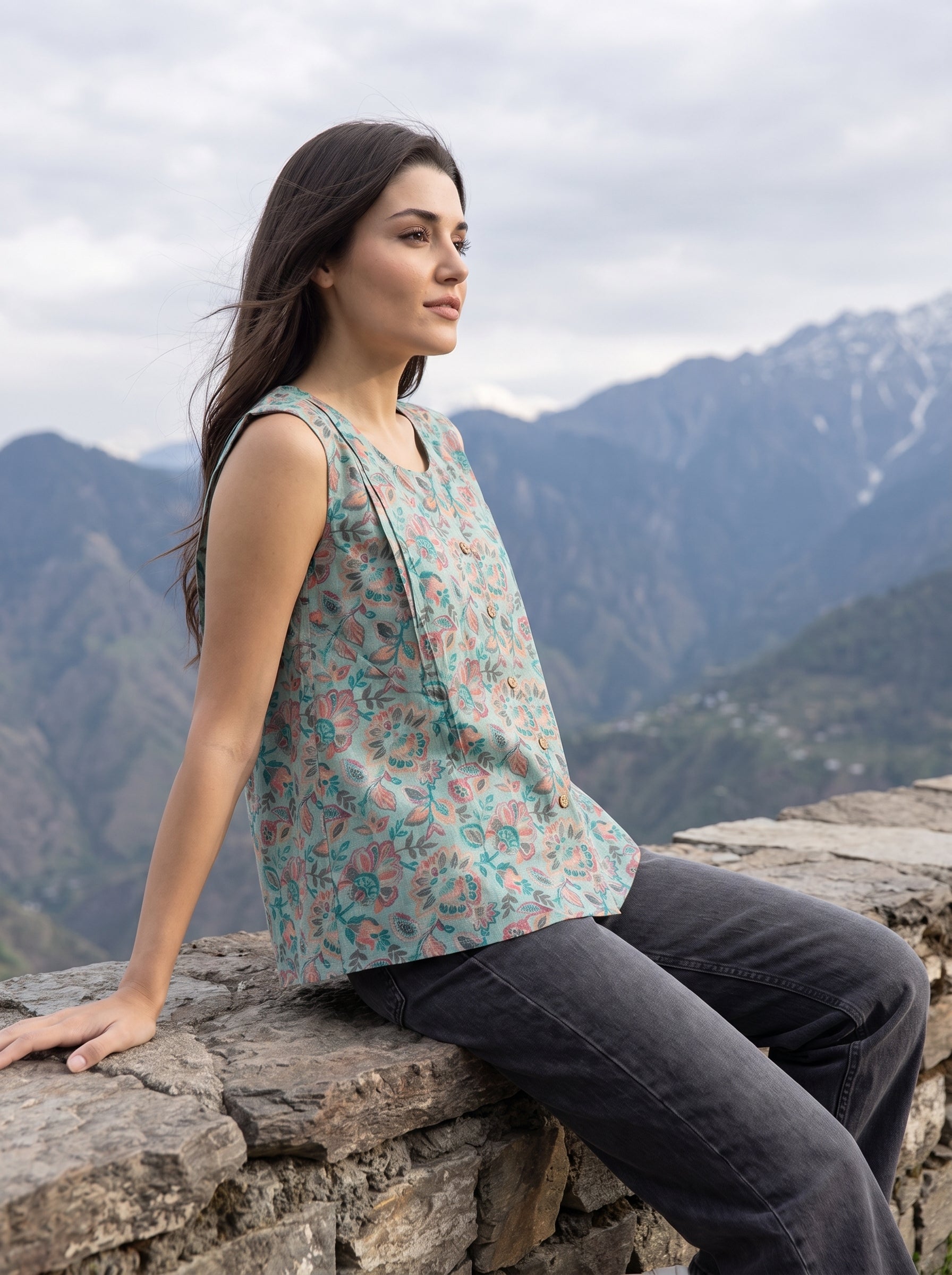 A woman sitting on a stone wall outdoors, wearing a sleeveless floral top with a teal, pink, and beige pattern, paired with dark gray jeans, with mountains in the background.