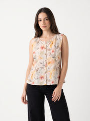 A woman modeling a sleeveless button-front top with a soft floral print in peach, orange, yellow, and muted green on a light beige background, paired with black pants against a plain light gray studio backdrop.