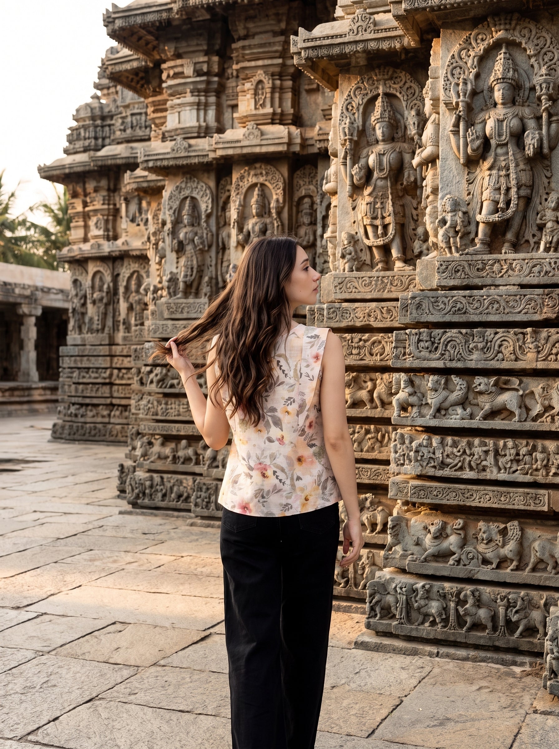 A woman stands in front of an ornate stone temple with detailed carvings, wearing a sleeveless cream top with a colorful floral print and black pants.