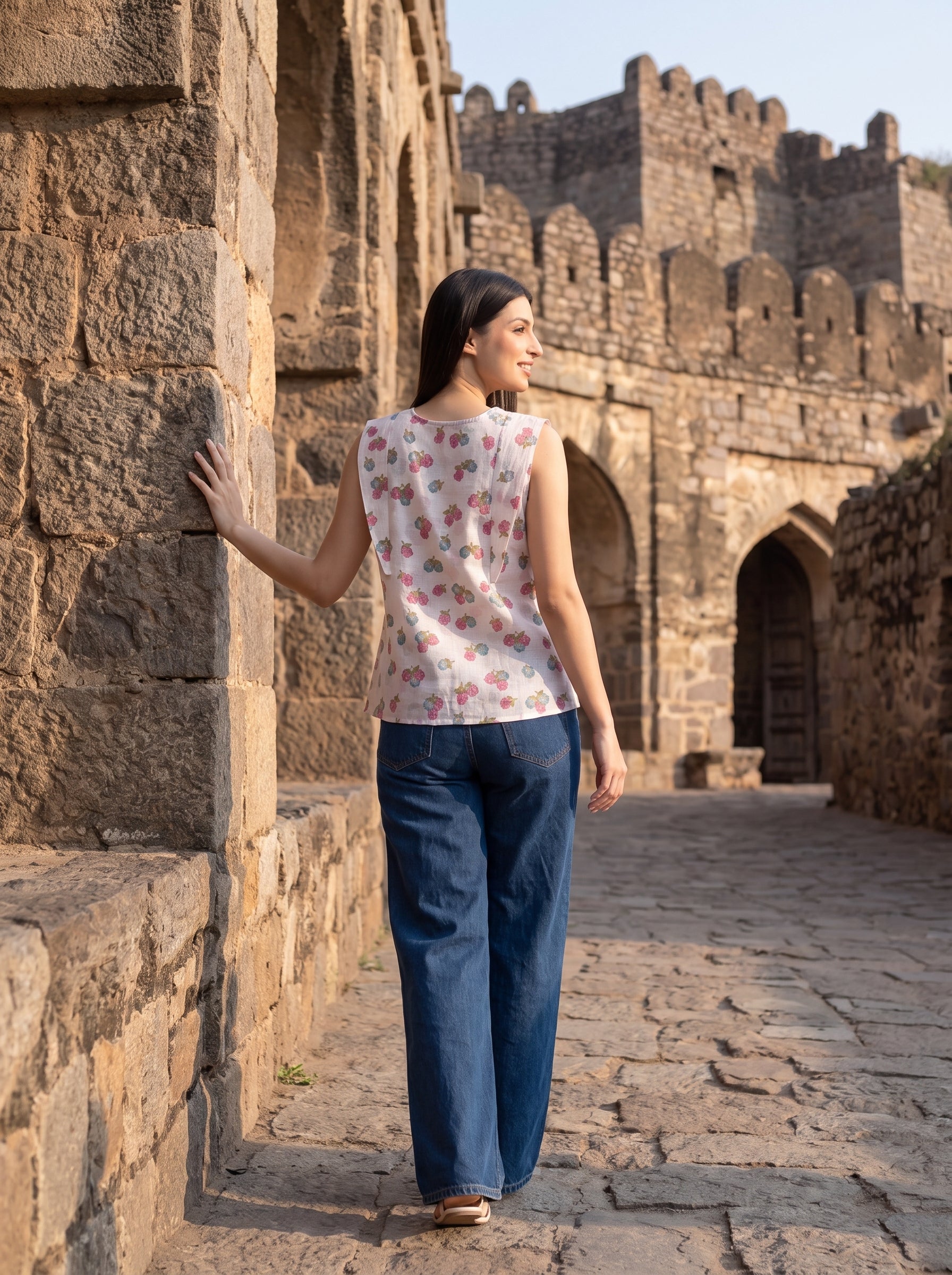 A woman wearing a sleeveless white top with a pink floral pattern and wide-leg blue jeans walks along a stone pathway beside ancient stone walls.