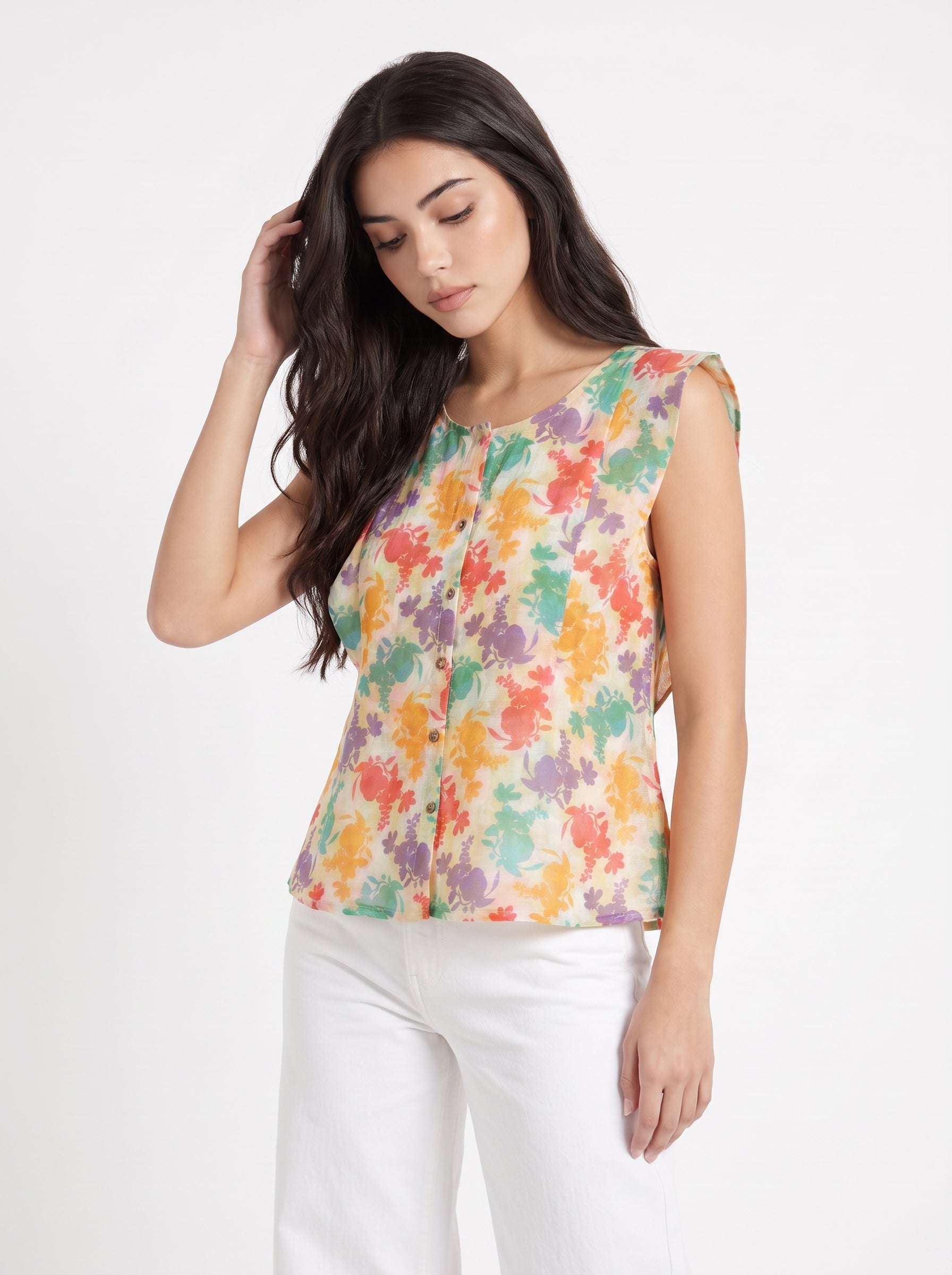 A woman wearing a sleeveless, button-front top with a colorful abstract floral print on a cream background, paired with white pants, photographed against a plain light gray studio background.