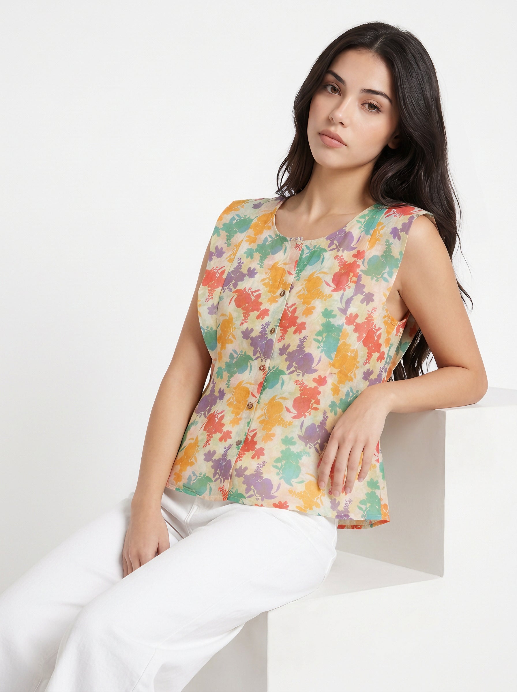 A woman wearing a sleeveless top with a colorful abstract floral print on a cream background, paired with white pants, sitting on a white geometric block against a white studio background.