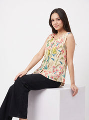A woman sits on a white block wearing a sleeveless beige blouse with a colorful floral and botanical print, paired with black wide-leg pants, against a plain white studio background.