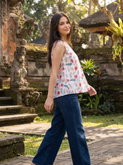 A woman wearing a sleeveless white floral top and dark blue wide-leg jeans walks outdoors in a tropical garden setting with stone structures and greenery.